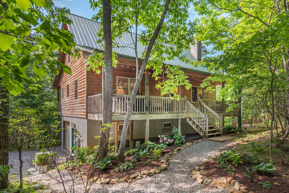 The cabin's exterior is surrounded by lush greenery, featuring a spacious porch with wooden railings. Steps lead up to the entrance, complemented by neatly arranged flower beds and gravel pathways, creating a welcoming approach among the trees.