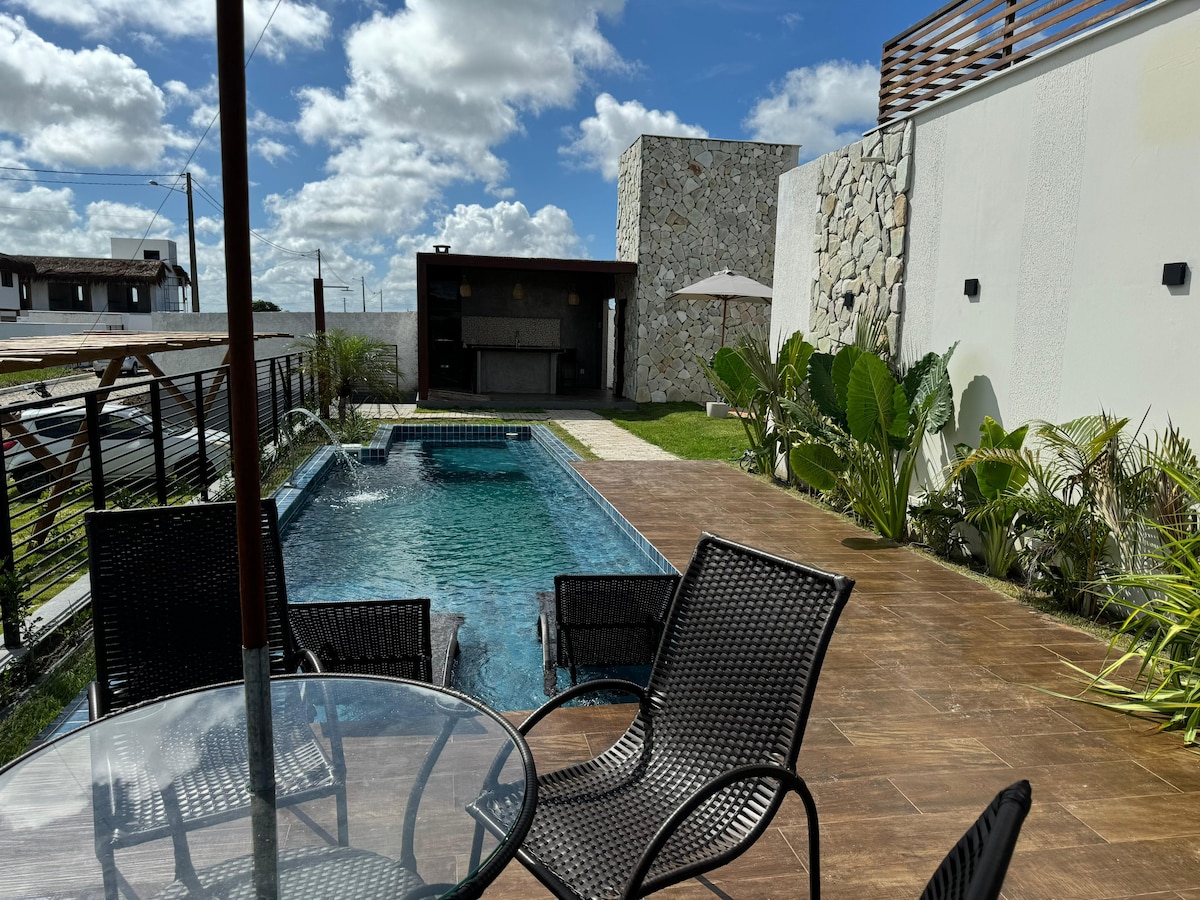 A sunlit outdoor area features a modern pool surrounded by a wooden deck. A glass table with black chairs is positioned nearby, complemented by lush green plants. A shaded lounge area with an umbrella provides a comfortable spot to relax.