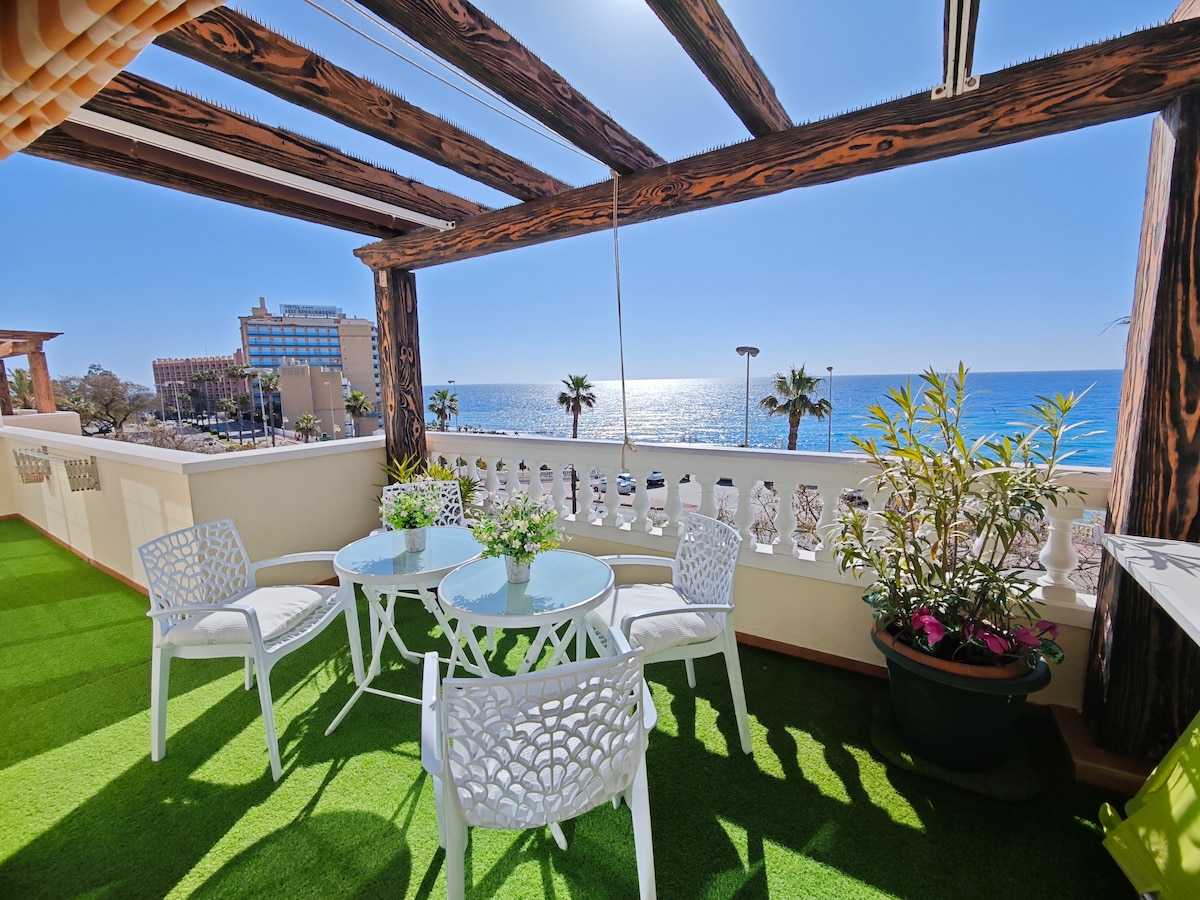 An outdoor terrace is displayed, featuring a glass-top table surrounded by four white chairs. Lush greenery decorates the space, along with potted plants. The stunning view of the ocean and clear blue sky adds a vibrant touch, creating an inviting area for relaxation.