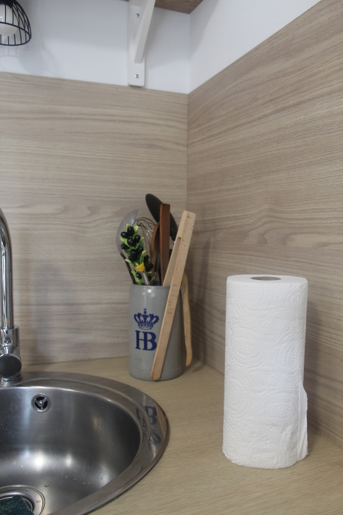 A well-equipped kitchen corner features a stainless steel sink with a modern faucet. Utensils are organized in a holder, complemented by a roll of paper towels for convenience. The warm wood tones of the cabinetry and countertop create an inviting cooking space.
