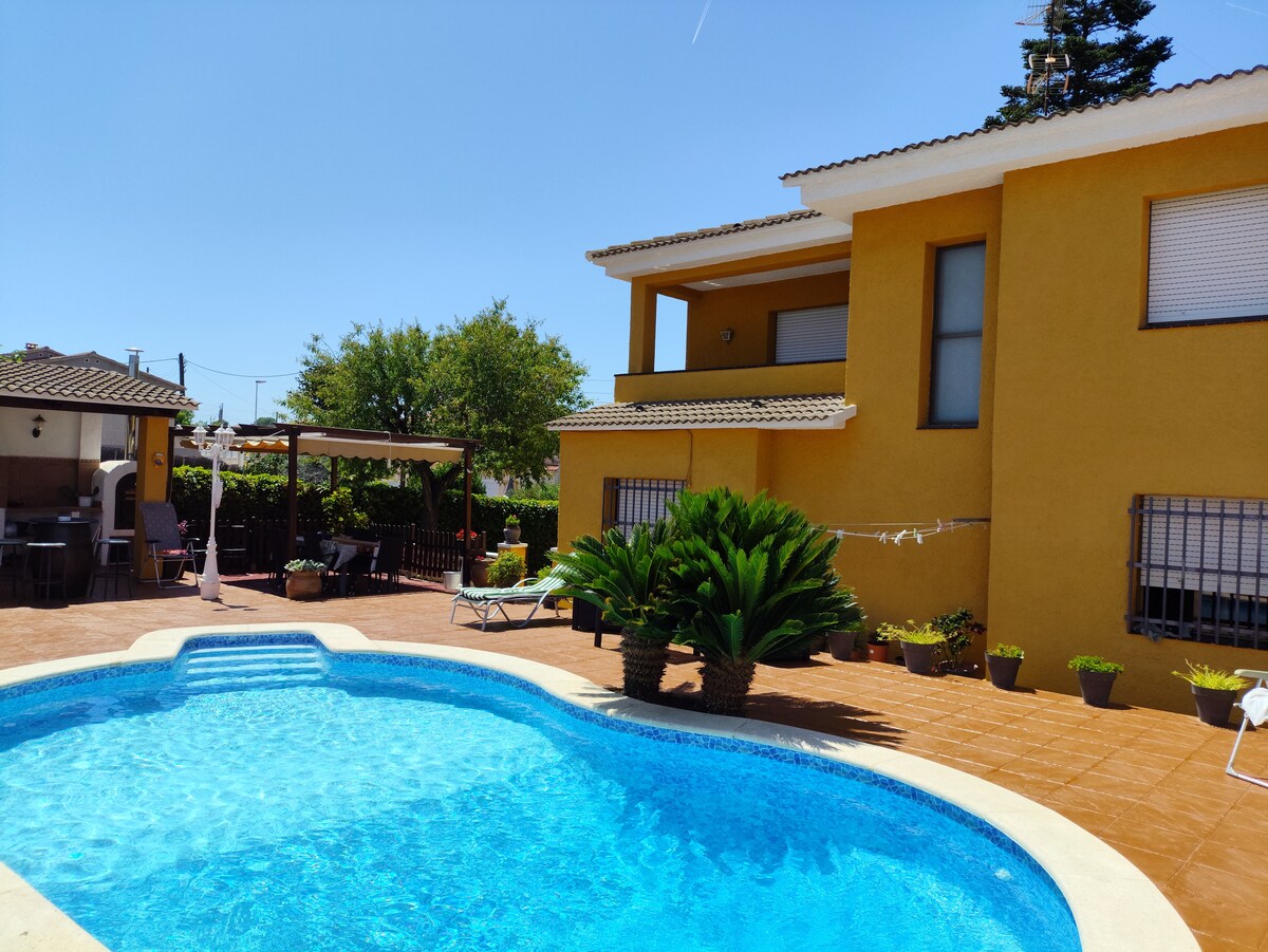 A vibrant yellow house features a welcoming outdoor area with a clear blue pool. A spacious patio surrounds the pool, complemented by tropical plants. Nearby, a shaded seating area with a pergola provides a relaxing spot, enhancing the outdoor atmosphere.