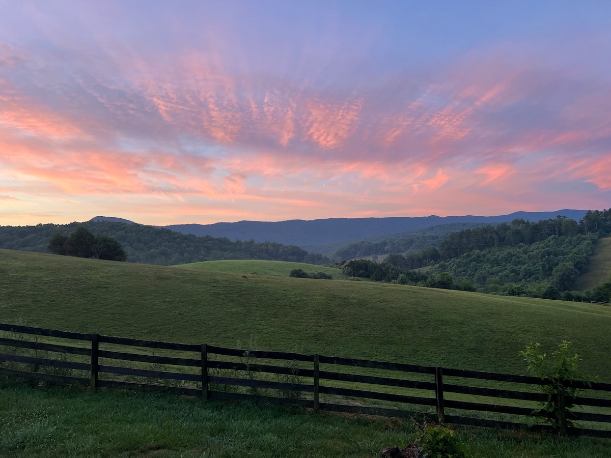 A panoramic view of rolling green hills is presented under a sunset sky, with soft pink and purple hues blending into light blue. A wooden fence lines the foreground, framing the serene landscape, while the distant mountains create a tranquil backdrop.