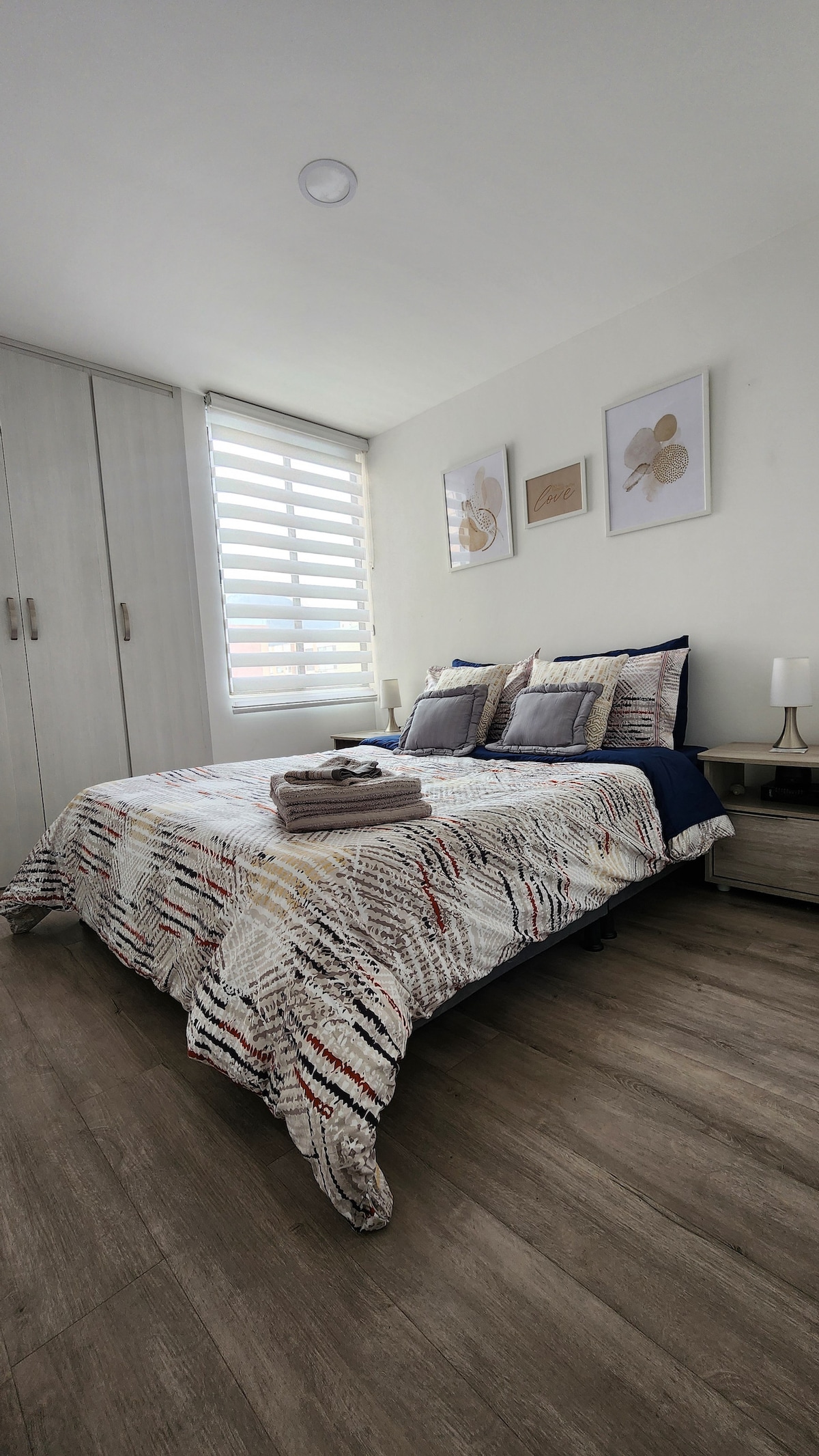 A spacious bedroom is presented, featuring a patterned bedspread on a large bed. Soft pillows are arranged at the head of the bed, with two nightstands on either side. Natural light filters through modern window treatments, highlighting the calming decor and wooden flooring.