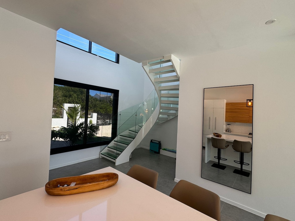 An airy interior showcases a modern spiral staircase with glass railings, leading to the upper level. Natural light floods the space through large windows, highlighting a stylish kitchen area with two bar stools and contemporary cabinetry, all accented by a neutral color palette.