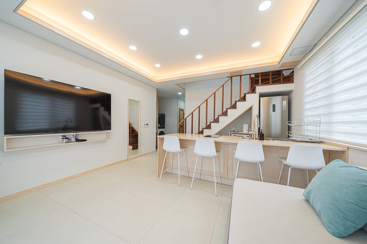 The open living area features modern furnishings, including a large television mounted on the wall and four white chairs positioned at a sleek kitchen counter. A staircase leads to the upper level, with soft lighting illuminating the space.