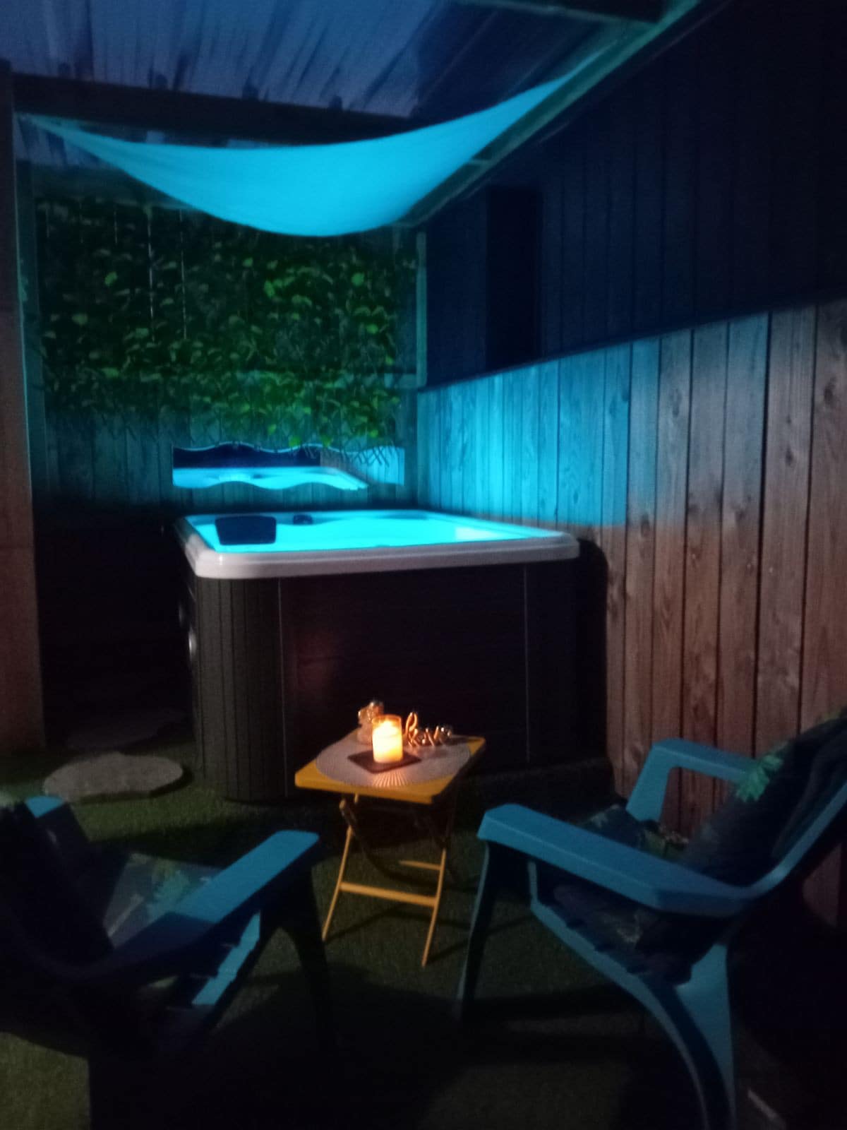 A private outdoor area features a well-lit hot tub, surrounded by wooden walls and greenery. Two relaxed chairs face a small table, which is adorned with a candle. Soft blue lighting creates a calming ambiance, enhancing the secluded space.