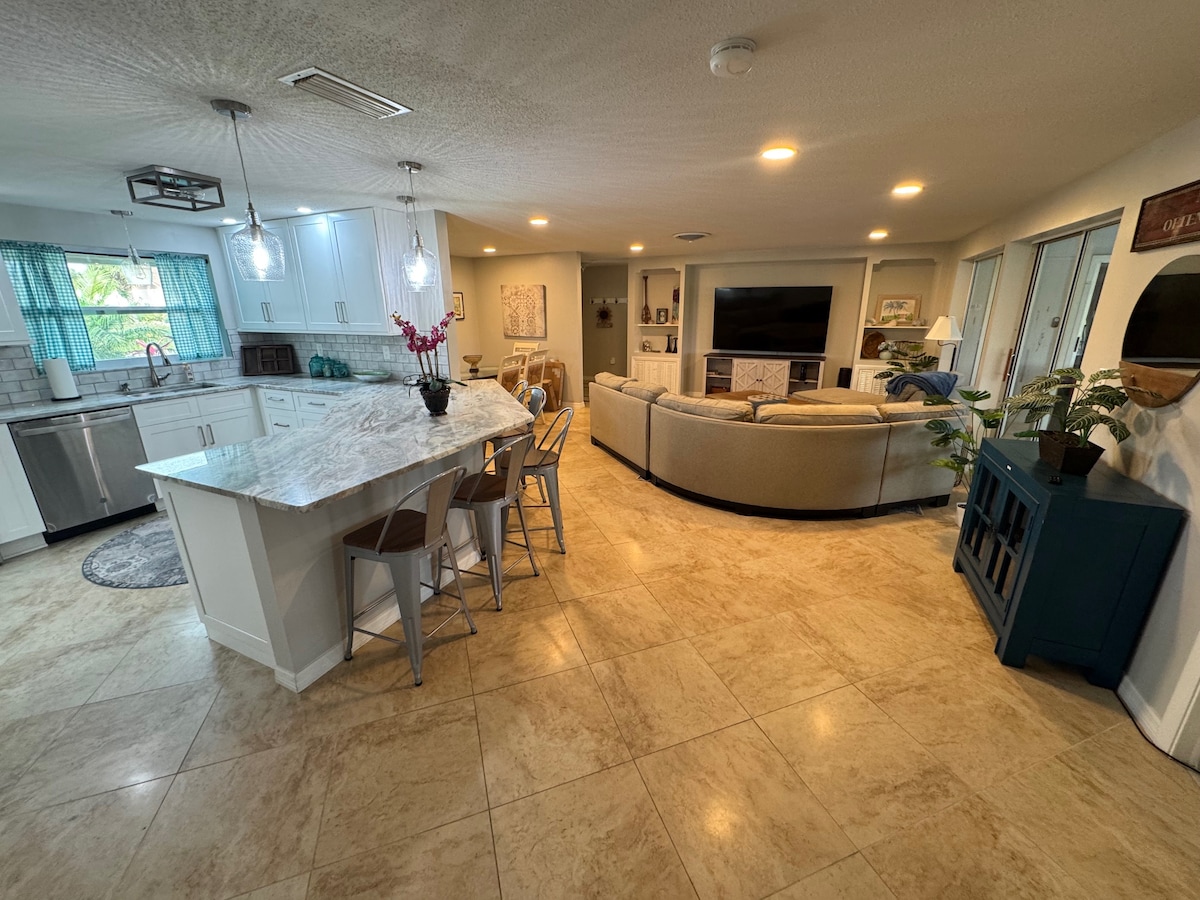 An open living space features a modern kitchen with a large island and stainless steel appliances. Adjacent is a cozy seating area with a sectional sofa, creating an inviting environment for social gatherings. Ample natural light streams through nearby windows, enhancing the room's brightness.