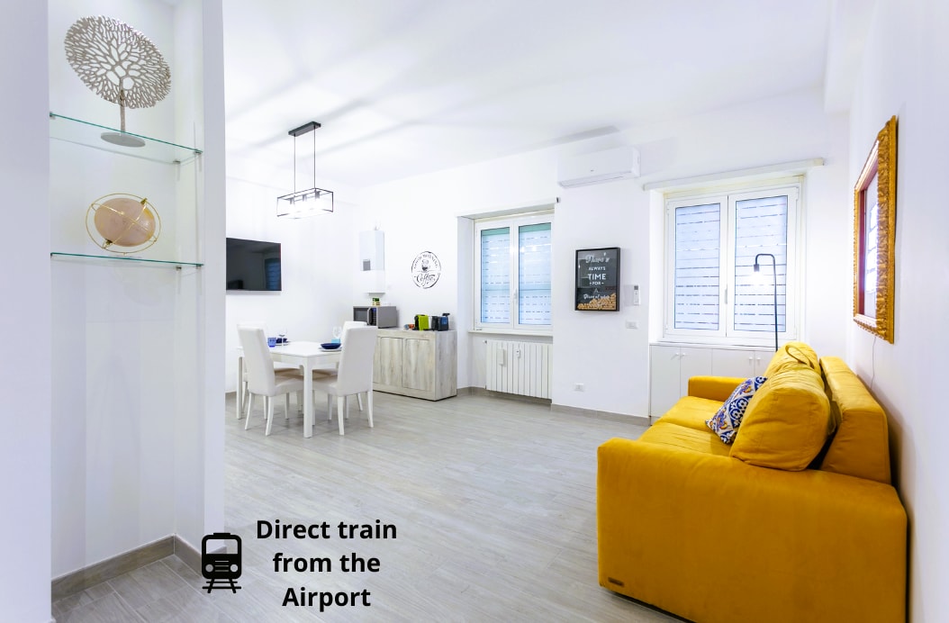 A spacious living area features a bright yellow sofa and a dining table set for four. Large windows provide natural light, with a Smart TV mounted on the wall. A small shelf showcases decorative items, contributing to a modern atmosphere.