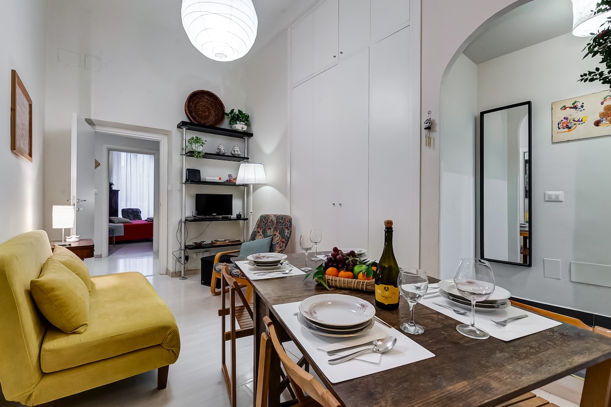 A dining area features a wooden table set with plates and glassware, accompanied by vibrant fruits in a basket. A yellow sofa adds a pop of color, while soft lighting is provided by a round ceiling fixture. A mirror and a cozy living space are visible in the background.