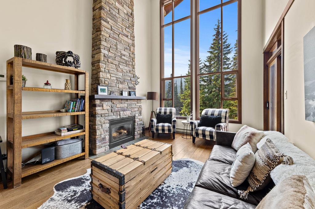 A spacious living area features a stone fireplace as a focal point. Two armchairs with checkered upholstery are positioned near the windows, offering views of surrounding trees. A wooden shelving unit displays books and decor, while a rustic coffee table sits on a patterned area rug.
