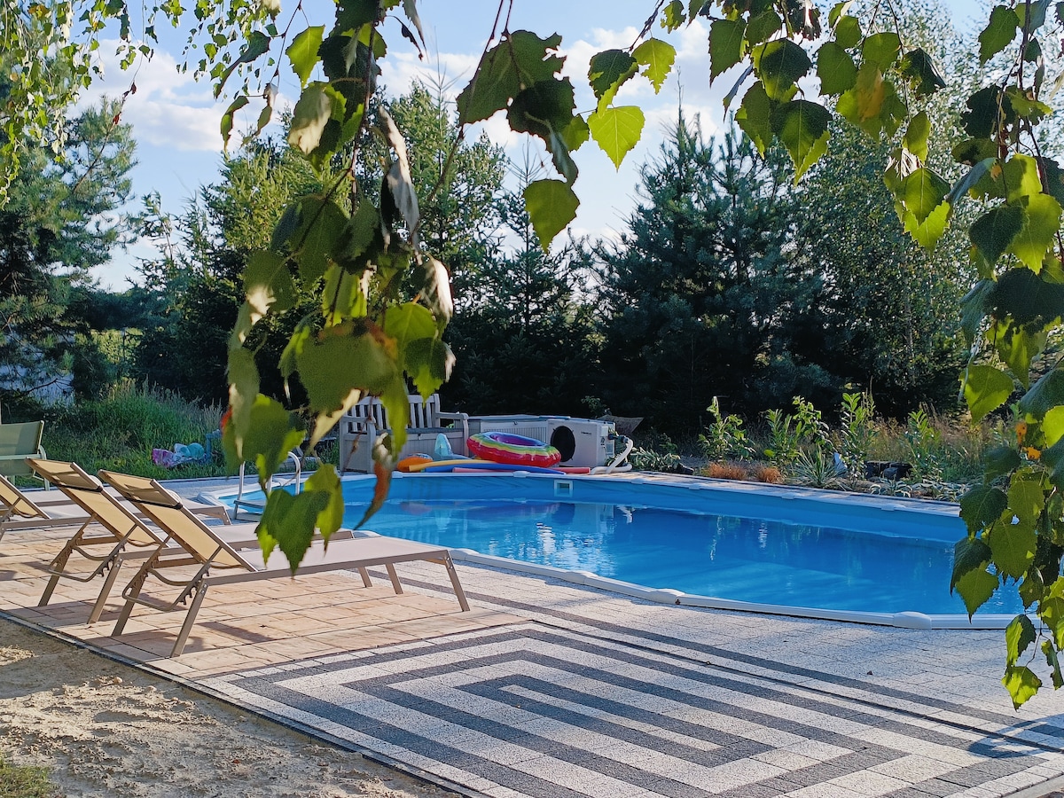 A rectangular swimming pool is framed by a decorative tile border and surrounded by lounge chairs. Lush greenery, including trees and shrubs, creates a natural backdrop, while colorful pool floats are visible nearby, adding a playful touch to the serene outdoor space.
