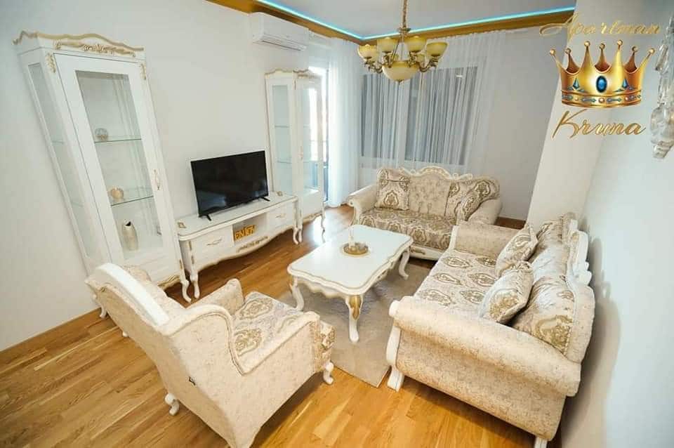 A well-appointed living area features elegant seating arrangements with upholstered sofas and armchairs. A central coffee table sits atop a light-toned area rug. A flat-screen television is mounted against a light wall, while natural light filters through sheer curtains adorned with a soft glow.