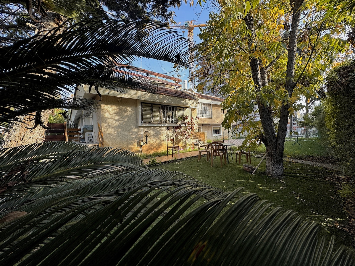 A view of the apartment's outdoor space is framed by lush greenery, showcasing a spacious garden area with a dining table and chairs. A large tree provides shade, and the two-story building features windows and a patio area, highlighting natural surroundings.