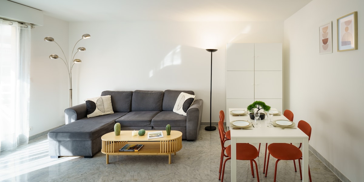 A bright living area features a spacious gray sofa complemented by a light wood coffee table. A dining table set for four is visible, surrounded by red chairs. A lamp provides ambient light, and a cabinet stands against the wall, enhancing the room's functionality.