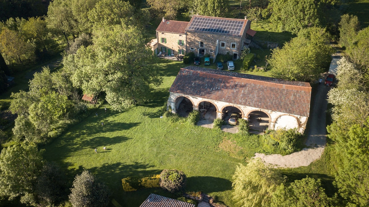 An aerial view captures the spacious property, surrounded by lush greenery and mature trees. A charming building with solar panels is visible alongside an arched structure. The well-maintained grounds feature open grassy areas and a pathway leading to the entrance.