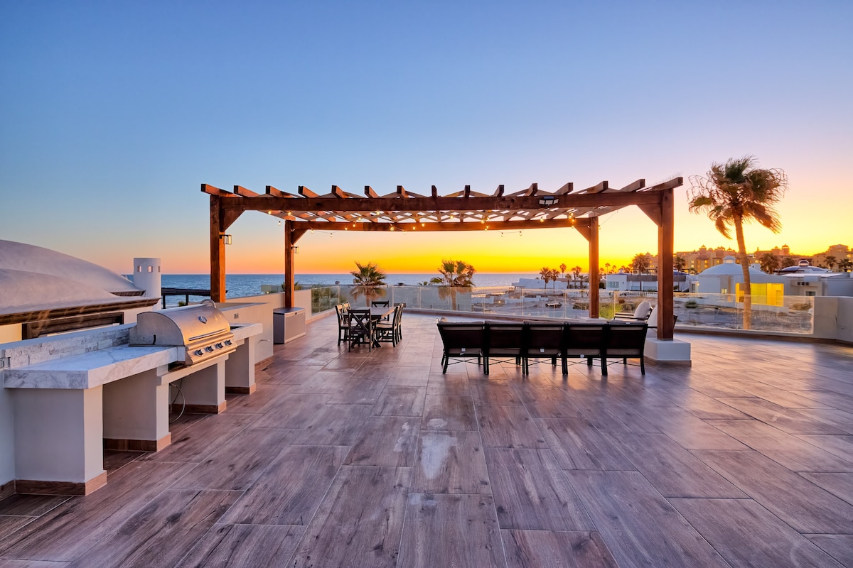 An expansive outdoor terrace features a wooden pergola, offering shade over a dining set and lounge area. A built-in barbecue station is positioned to one side, while stunning ocean views and a colorful sunset are visible in the background, enhancing the outdoor living space.