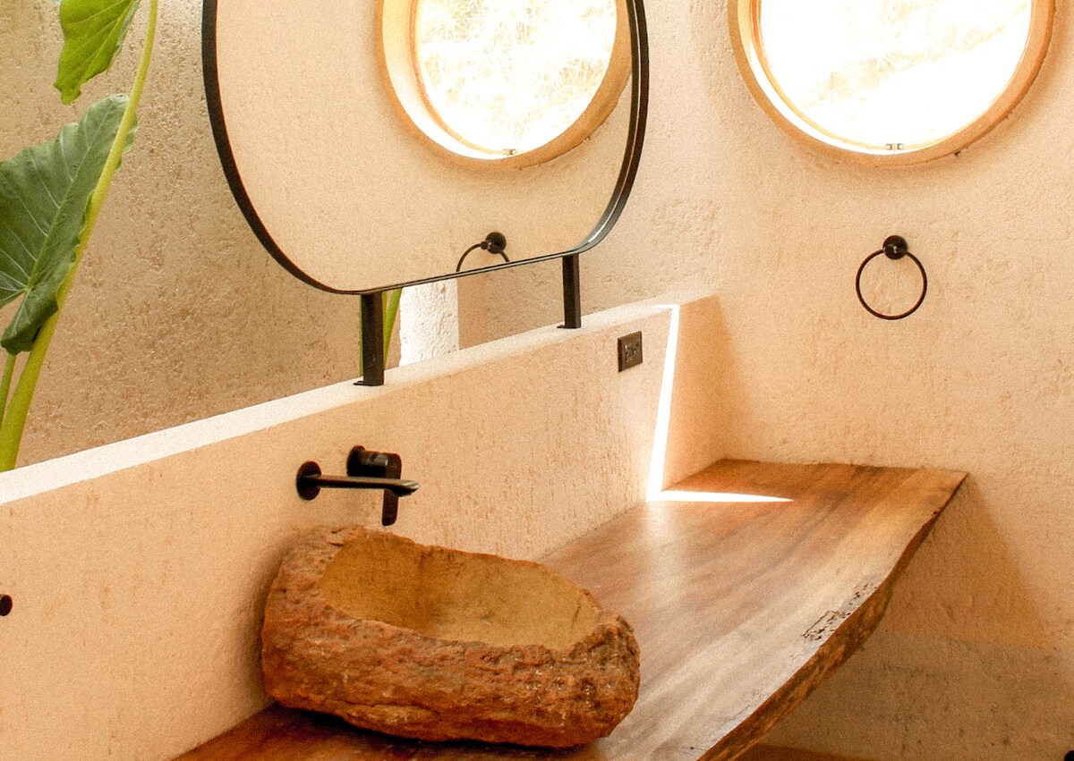 A well-lit bathroom space features a wooden countertop with a natural stone basin. Two circular mirrors reflect natural light through round windows, while minimalistic fixtures enhance the serene atmosphere. Green foliage adds a touch of nature to the inviting environment.
