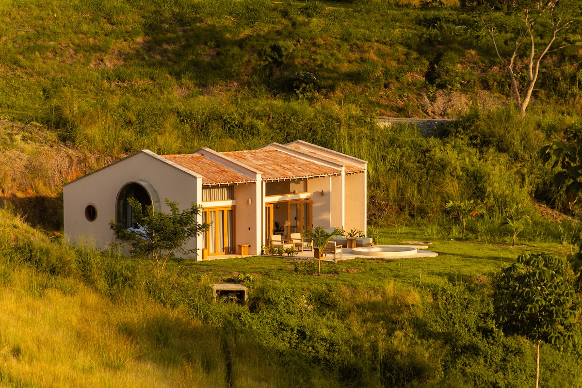 A charming villa is nestled in a lush hillside, featuring a welcoming facade with a sloped roof. Large windows provide views of the surrounding greenery and access to a private patio with a small pool, ideal for relaxation in a serene environment.