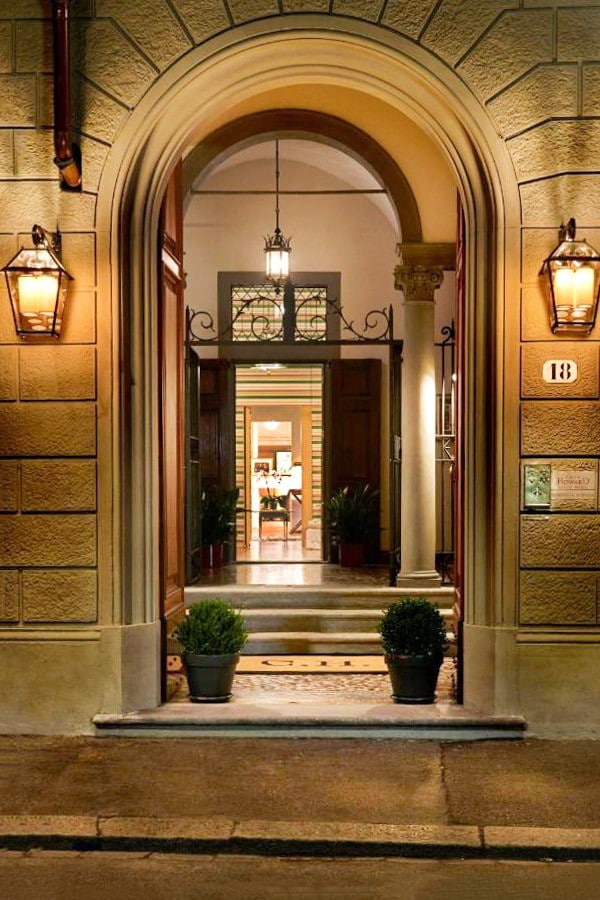 The entrance of the building features an elegant archway framed by polished stone. Two lanterns illuminate the doorway, leading to a glimpse of the welcoming interior. Potted plants enhance the inviting appearance, and the pathway is lined with cobblestones.