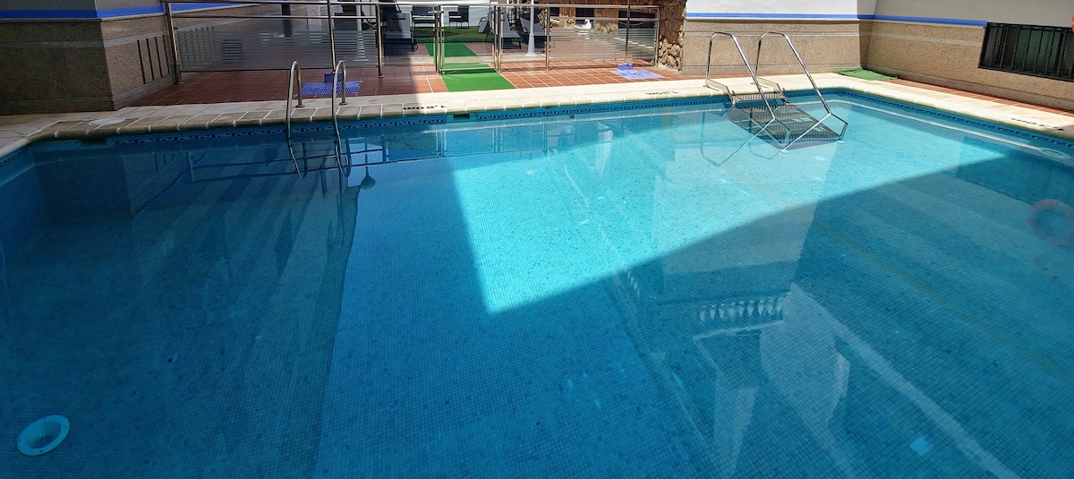 A clear and inviting salt-water pool is pictured, featuring a blue mosaic tile bottom. Two steps lead into the shallow end, while the surrounding area includes a spacious deck. Strong sunlight creates a reflective pattern on the water's surface, enhancing the tranquil atmosphere.
