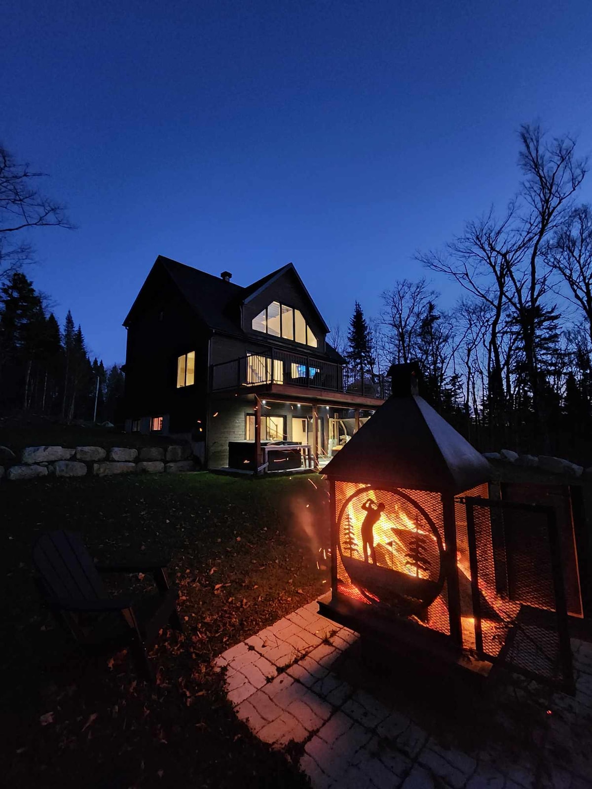 The exterior of the chalet is illuminated at dusk, showcasing a well-structured house with large windows. A fire pit is visible in the foreground, with flames flickering, providing warmth and ambiance. Surrounding trees stand silhouetted against the evening sky.