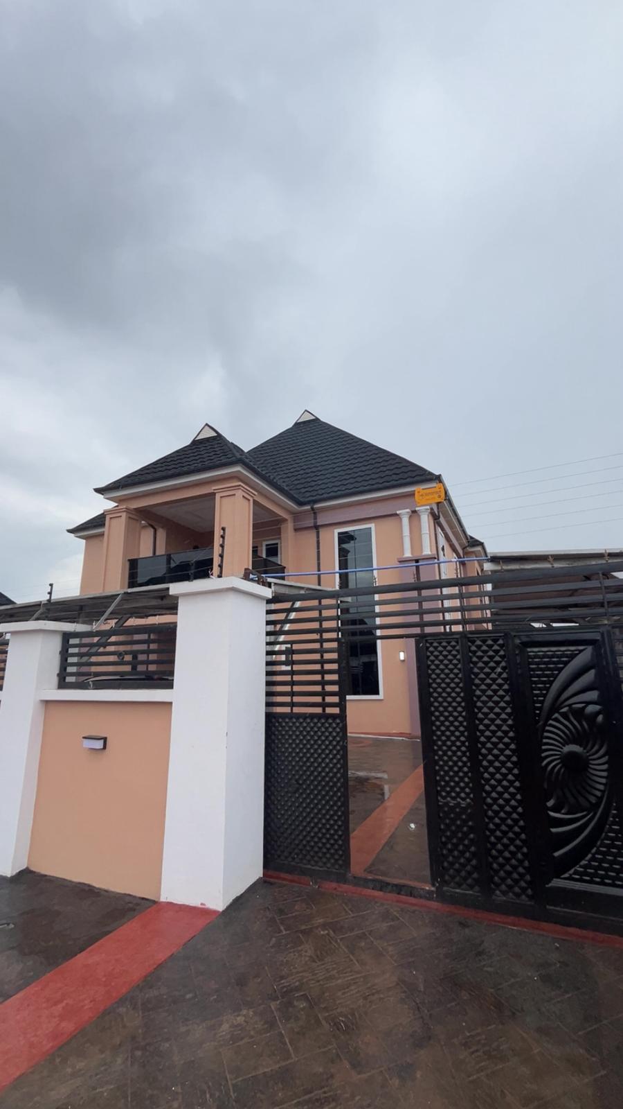 A modern, two-story house features a peaked roof and a blend of beige and pink exterior walls. A gated entrance is visible in the foreground, complemented by a black decorative fence. The sky appears overcast, creating a subdued atmosphere.