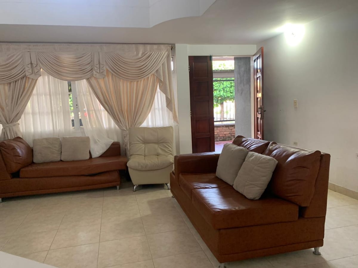 A spacious living area features two brown sofas with cushions, arranged to encourage conversation. Soft natural light filters through large windows adorned with elegant drapes. A decorative entrance reveals a glimpse of the outside, enhancing the welcoming ambiance of the room.