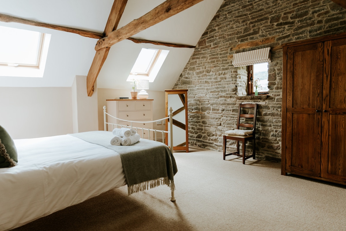 The spacious Master bedroom features a Superking bed with crisp linen and decorative towels neatly arranged. Natural light enters through skylights, illuminating the stone wall and wooden beams. A wooden wardrobe and a chair with a mirror complement the serene decor.