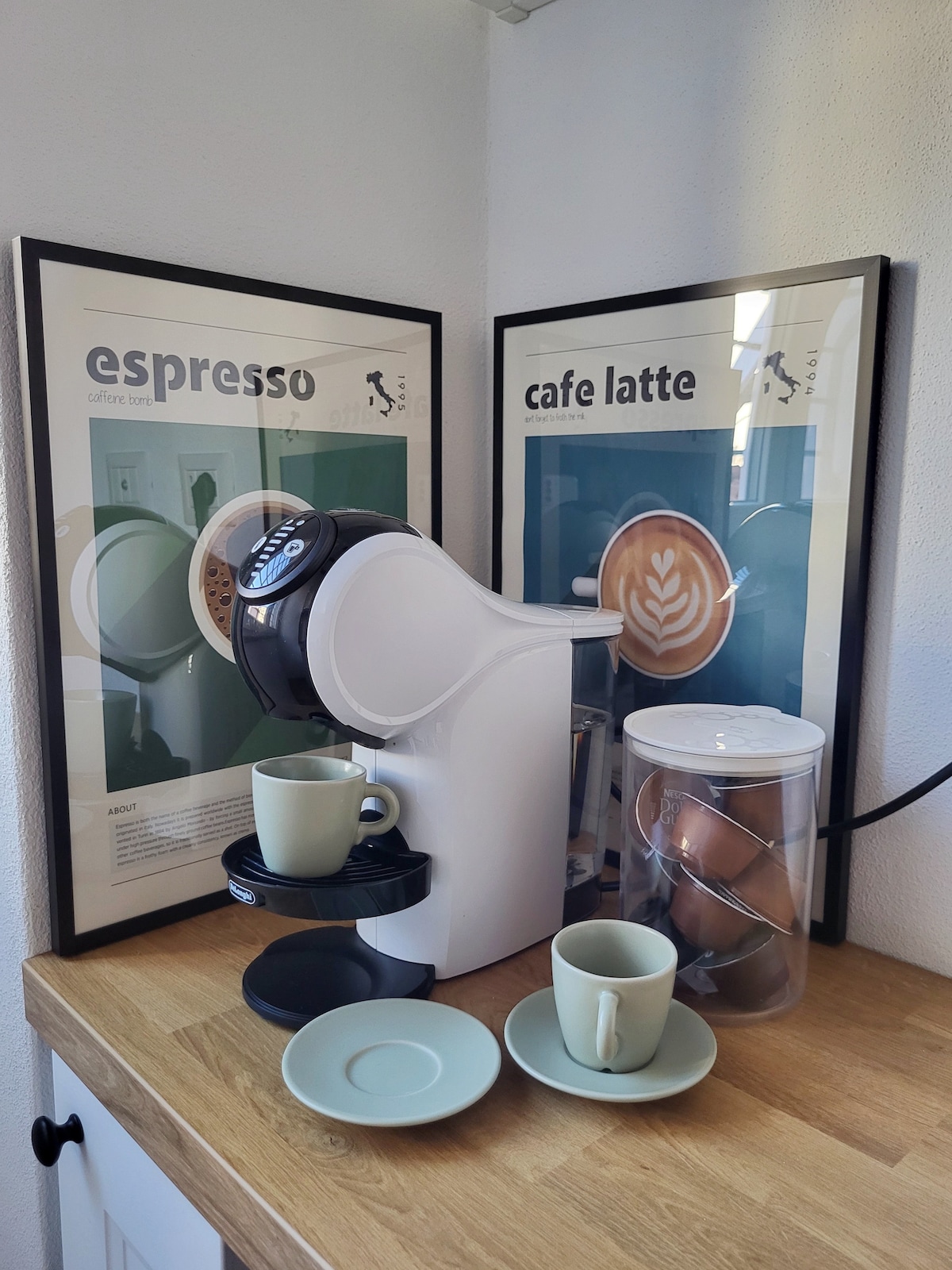 A modern coffee station showcases a coffee machine alongside two light green cups and matching saucers. Colorful coffee-themed artwork adorns the walls behind. The setup is located on a wooden surface, enhancing the cozy ambiance of the kitchen area.