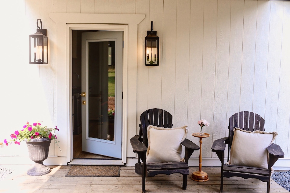 A welcoming porch area features two black wooden chairs with light cushions flanking a small, round table. A lantern hangs beside the door, and vibrant pink flowers bloom in a nearby planter. The open door leads into the interior space.