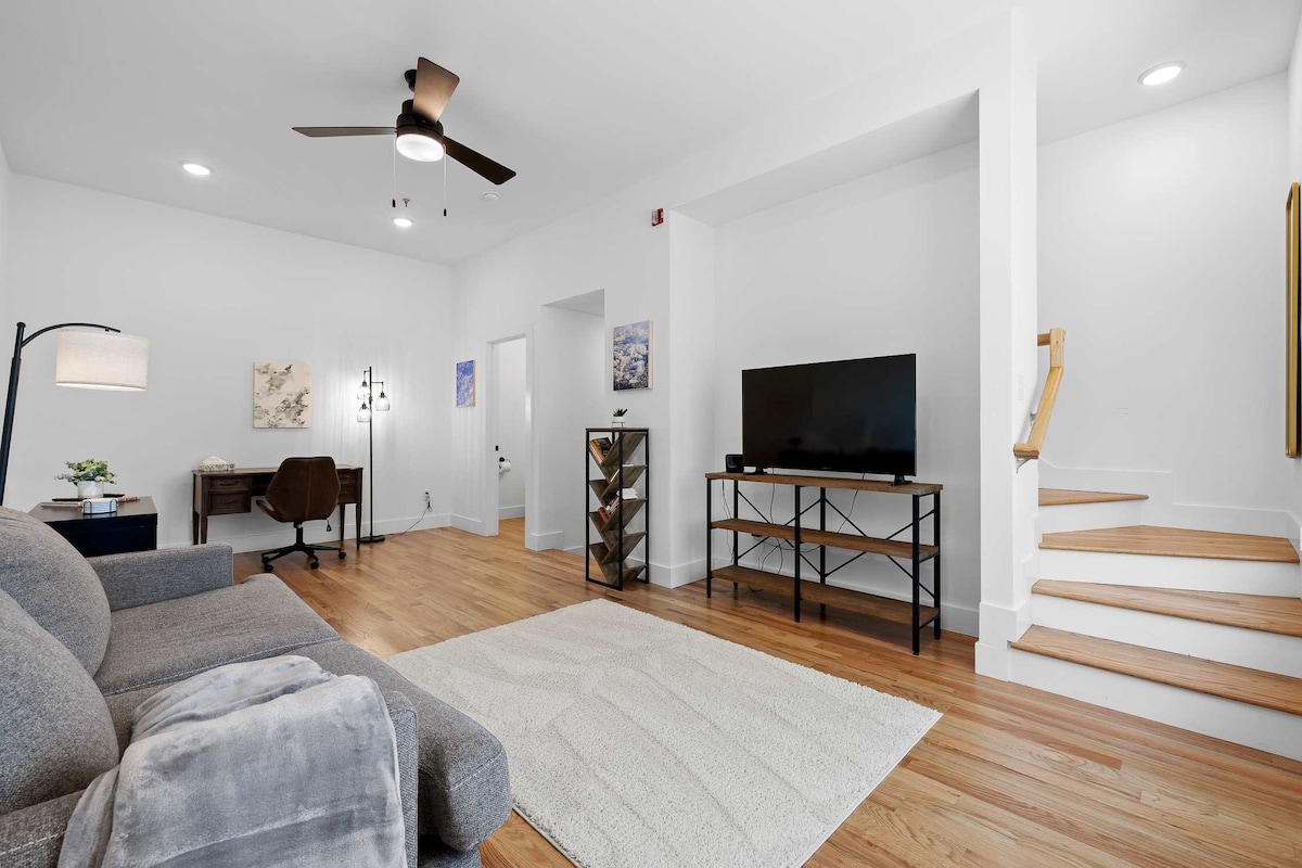 A bright living area features a comfortable gray couch and a sleek flat-screen TV mounted on a black console. A small desk is positioned nearby, and light wood flooring adds warmth. A staircase leads to the upper floors, with a decorative rug placed centrally on the floor.