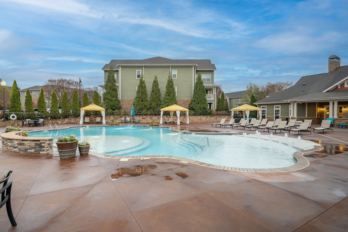 A resort-style pool area features a winding shape with shallow entry and lounge chairs surrounding it. Umbrellas provide shaded seating, and landscaped greenery lines the background. A spacious clubhouse is visible to the right, completing the inviting outdoor space.
