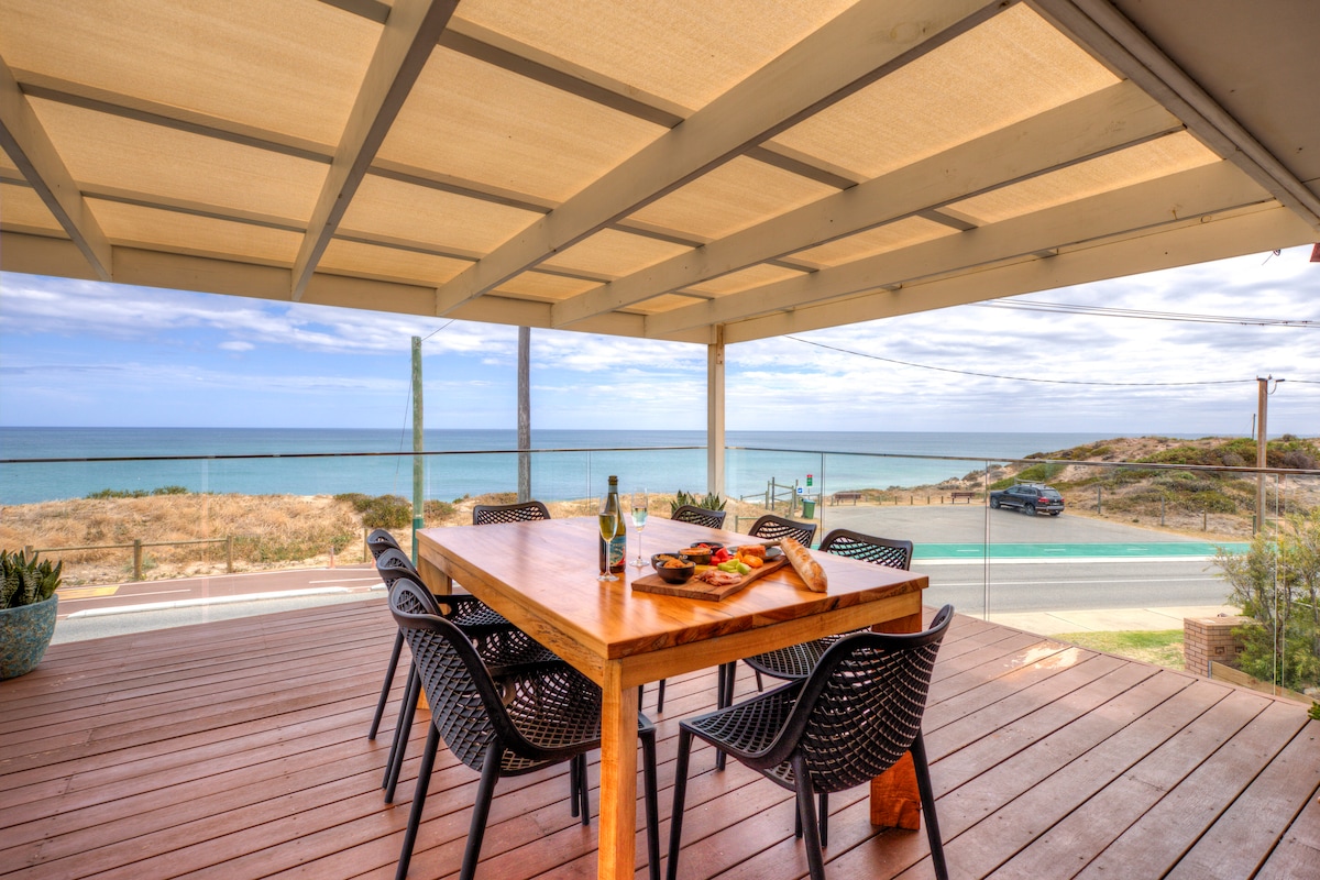 An expansive outdoor dining area features a large wooden table surrounded by six black woven chairs. The space is shaded by a covered roof, offering panoramic views of the ocean and beach beyond, with light wooden decking underfoot.