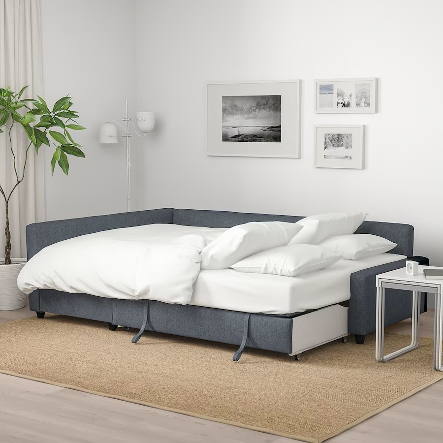 A comfortable sectional sofa is displayed, featuring a pull-out bed with a layered white comforter and multiple pillows. A small side table is placed beside the sofa, and a potted plant adds a touch of greenery to the cozy corner. Black-and-white artwork decorates the wall.