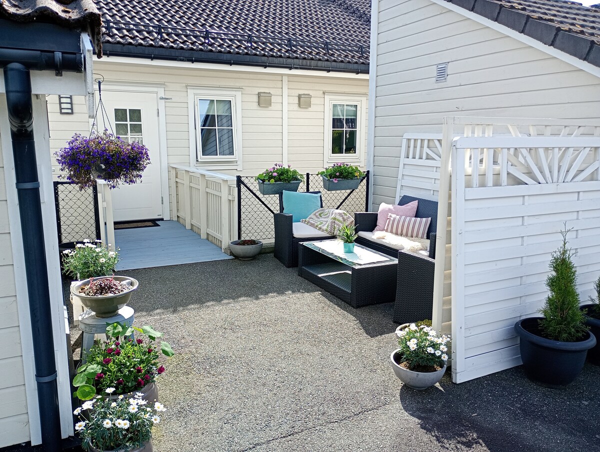 A private terrace area is presented, featuring a comfortable seating arrangement with light cushions. Potted plants and flowers are arranged around the space, adding greenery. The entrance door is visible, along with light-colored fencing that offers a sense of enclosure.