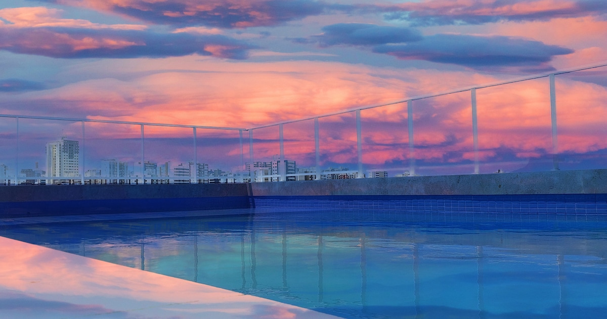 The rooftop pool area showcases a serene water surface reflecting a vibrant sunset sky, with clouds in shades of pink and purple. The infinity edge creates a seamless transition to the urban skyline, enhancing the tranquil ambiance of the space.