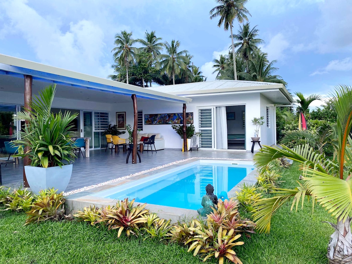 The image features a light-colored modern home with a private pool surrounded by a lush garden. Palm trees provide shade, and outdoor seating areas are visible. The pool area is framed by neatly arranged tropical plants, contributing to a serene outdoor setting.
