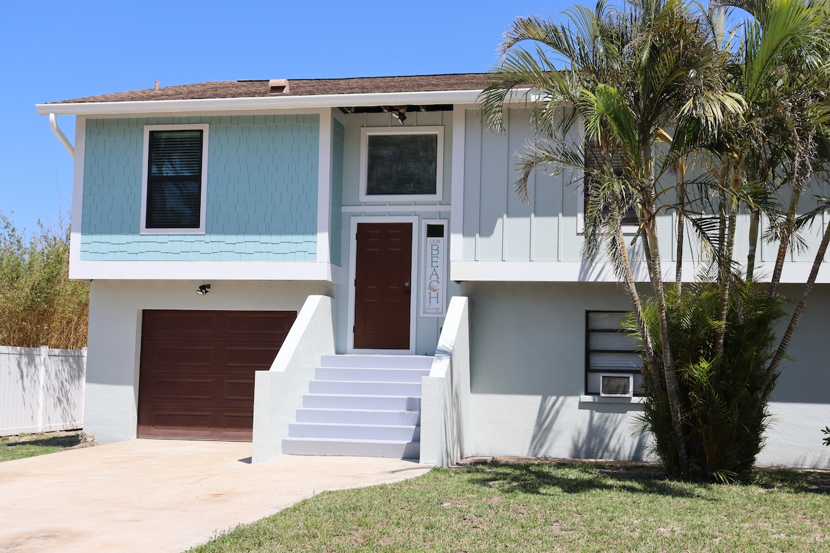A two-story house is shown with a light blue and white exterior. The entrance features a brown door with a decorative sign above it. Stairs lead up to the front door, and palm trees are positioned beside the driveway, providing a tropical touch.