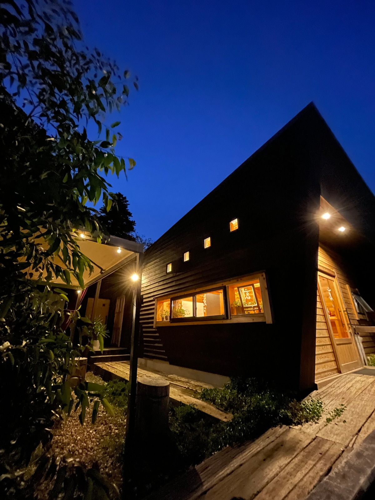 The exterior of the accommodation is showcased at twilight, with warm light illuminating the wooden structure. The angular roof design is complemented by multiple small windows, creating an inviting entrance framed by greenery. A wooden pathway leads towards the entryway.