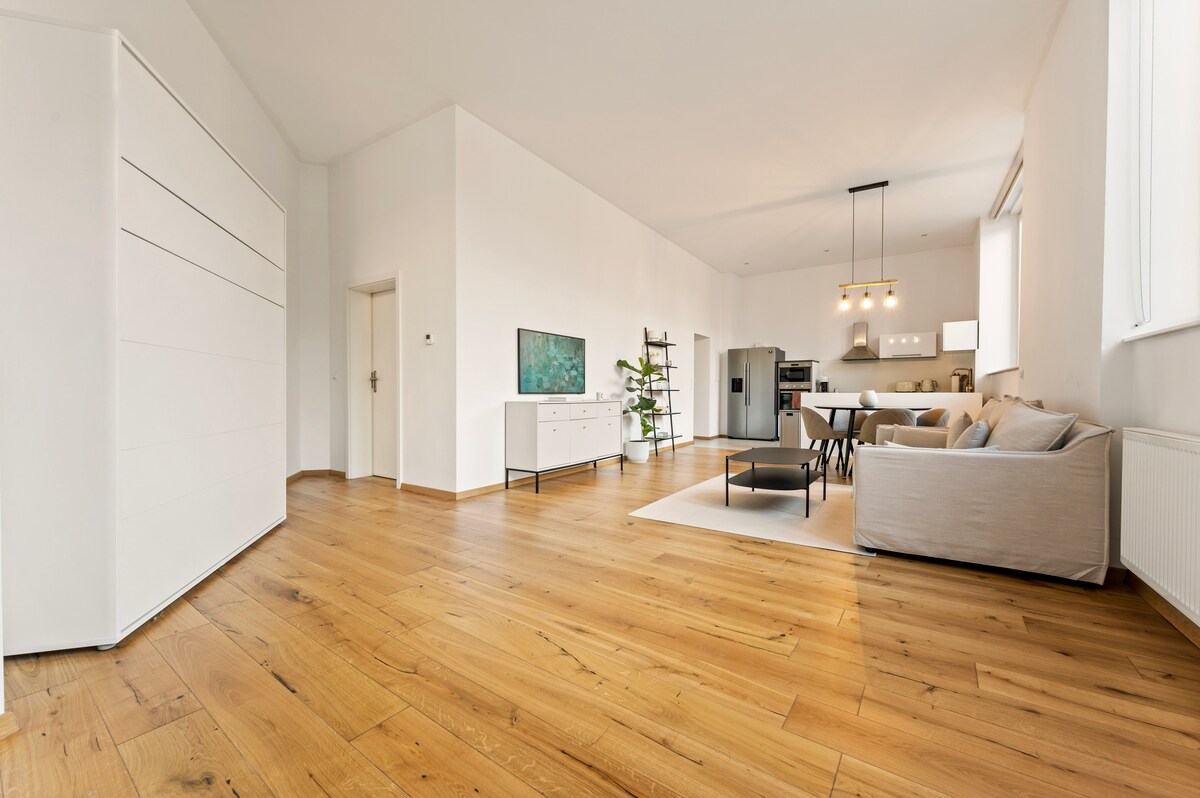 A spacious living area is presented with light wood flooring and white walls. A comfortable sofa faces a smart TV mounted on the wall. A modern dining table, surrounded by chairs, is located near a well-equipped kitchen, enhancing the open layout.