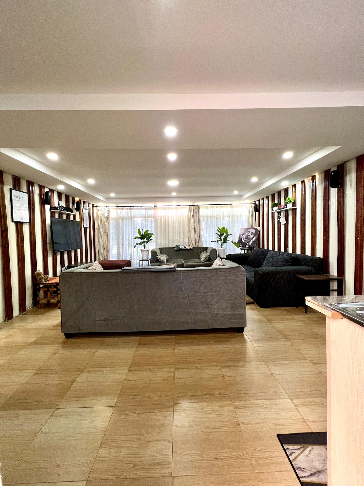 A spacious lounge area features soft seating arrangements with a mix of dark and light tones. Natural light filters through sheer curtains, illuminating the room. Decoratively arranged plants add a touch of greenery, while artistic wall accents bring character to the space.