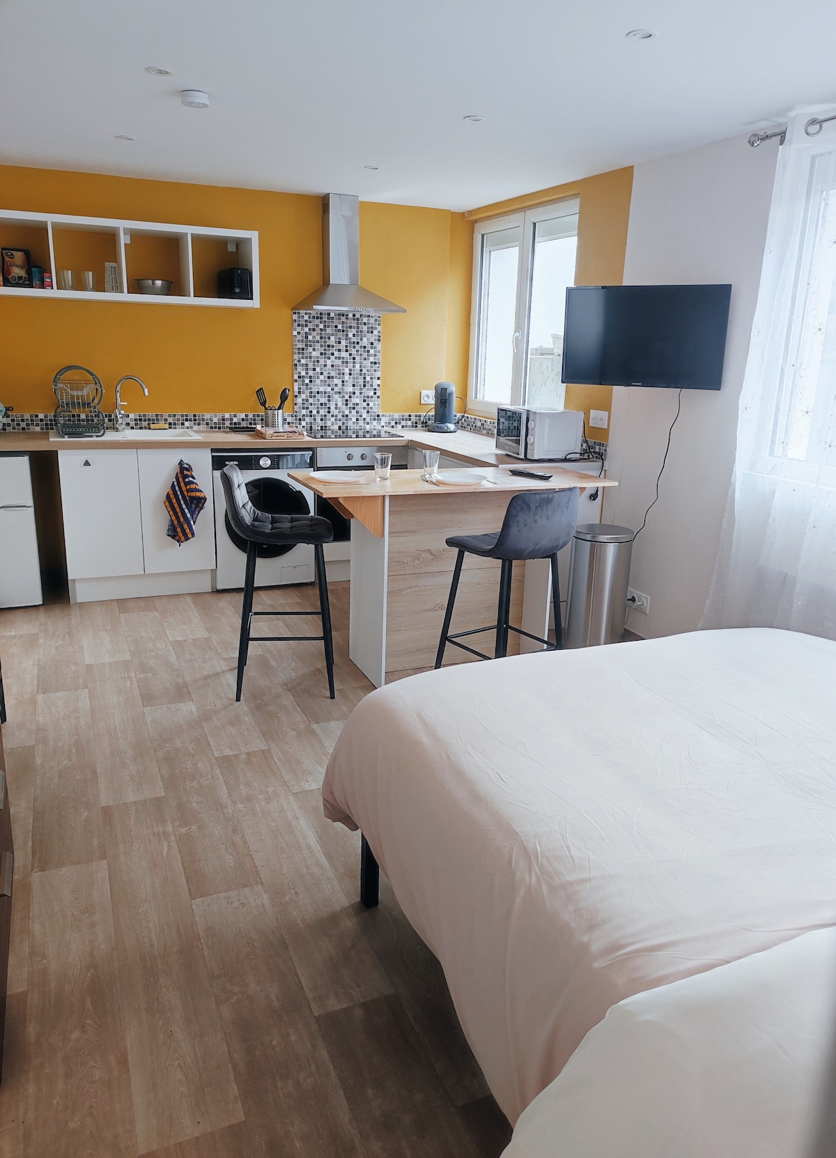 The studio features a well-equipped kitchen area with bright yellow walls. A spacious bed is positioned in the foreground, while a dining table with two stools and a wall-mounted TV are visible. Natural light filters through large windows, enhancing the open layout.