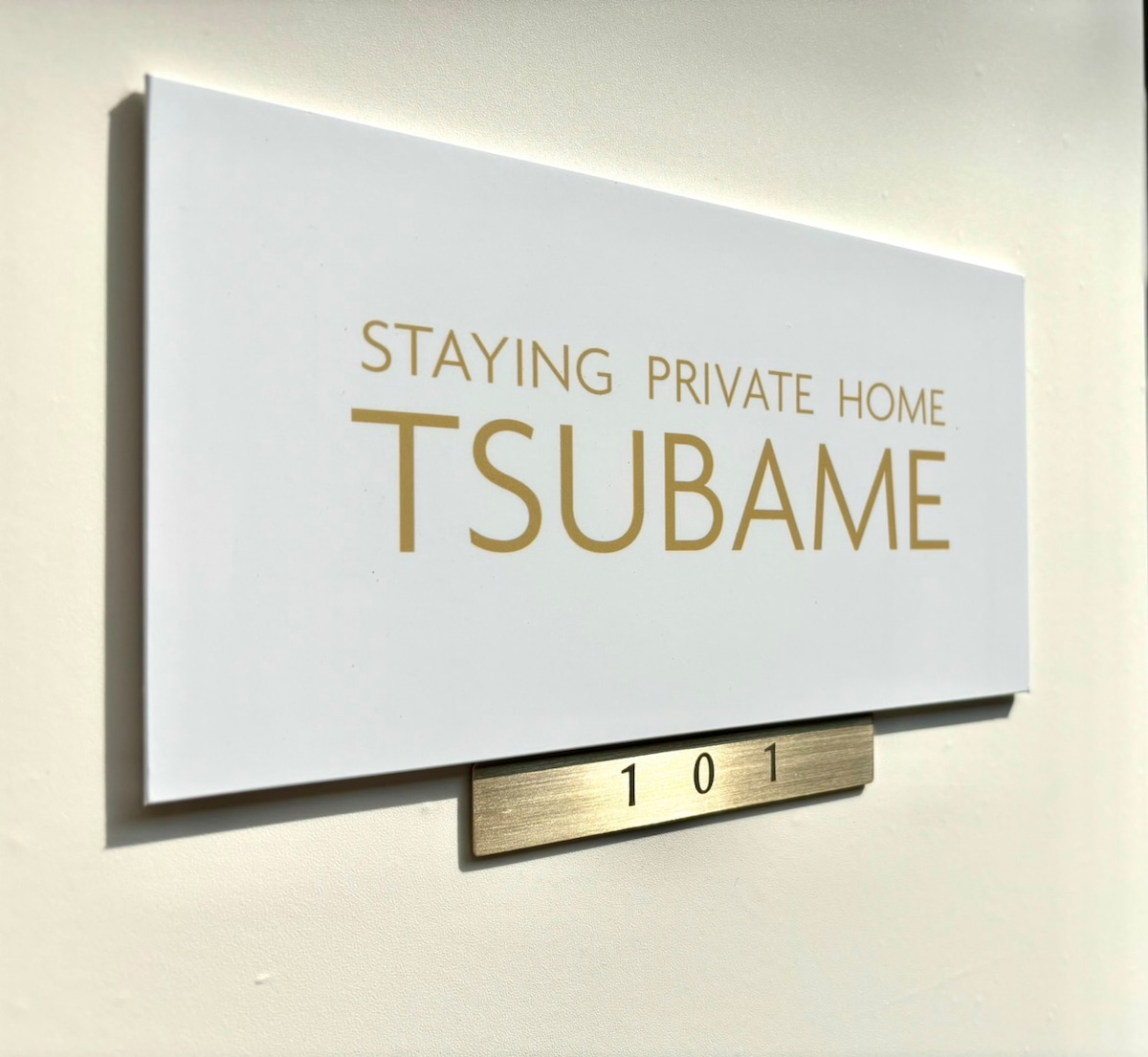 A wall-mounted sign indicates the property name, 'Staying Private Home Tsubame,' in elegant gold lettering. The number '101' is displayed below on a metallic plaque, contributing to a welcoming entryway.