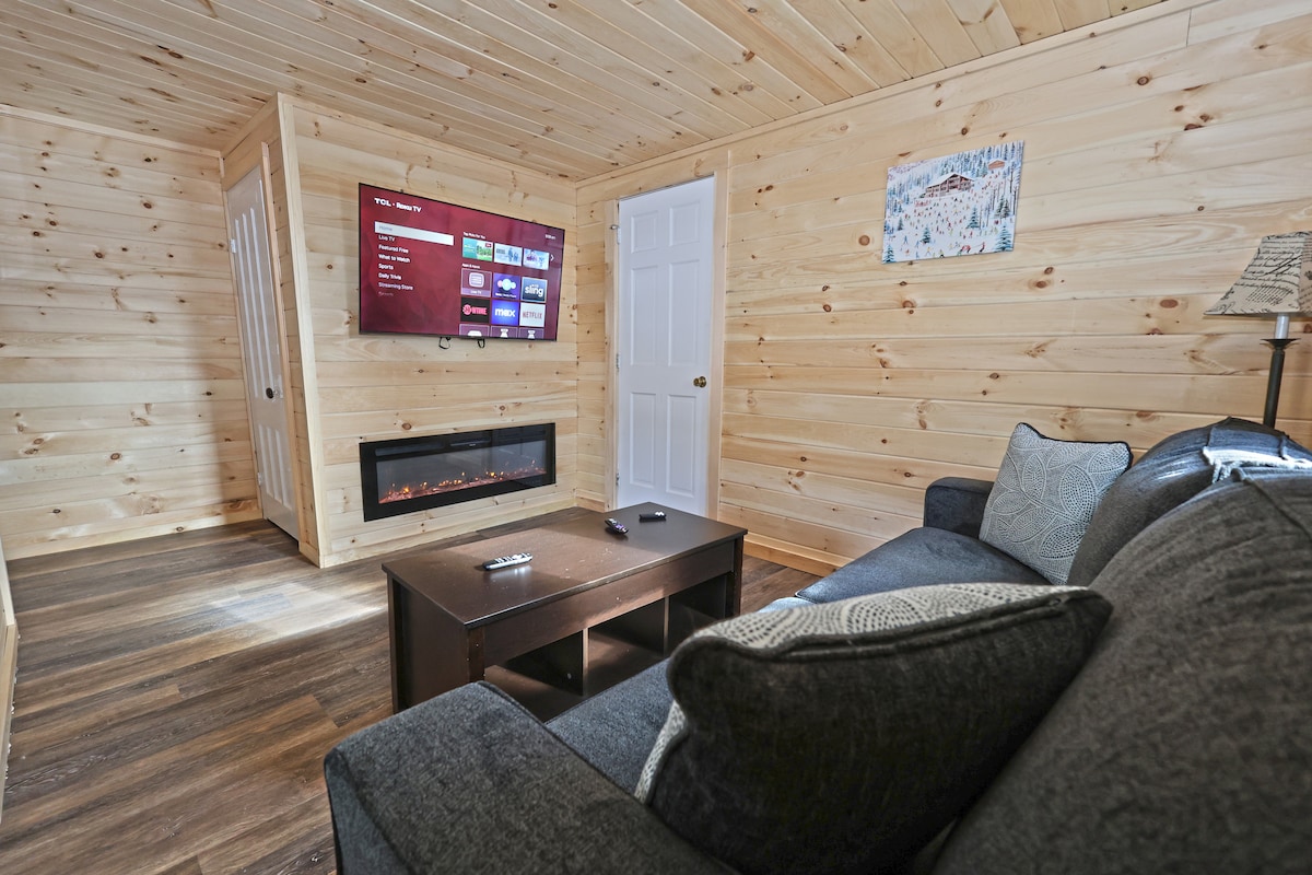 A cozy living area features a black sectional couch with decorative pillows, positioned across from a wall-mounted television. Below, an electric fireplace is integrated into the wooden wall paneling. Natural light filters in through a nearby door, illuminating the warm wood tones of the room.