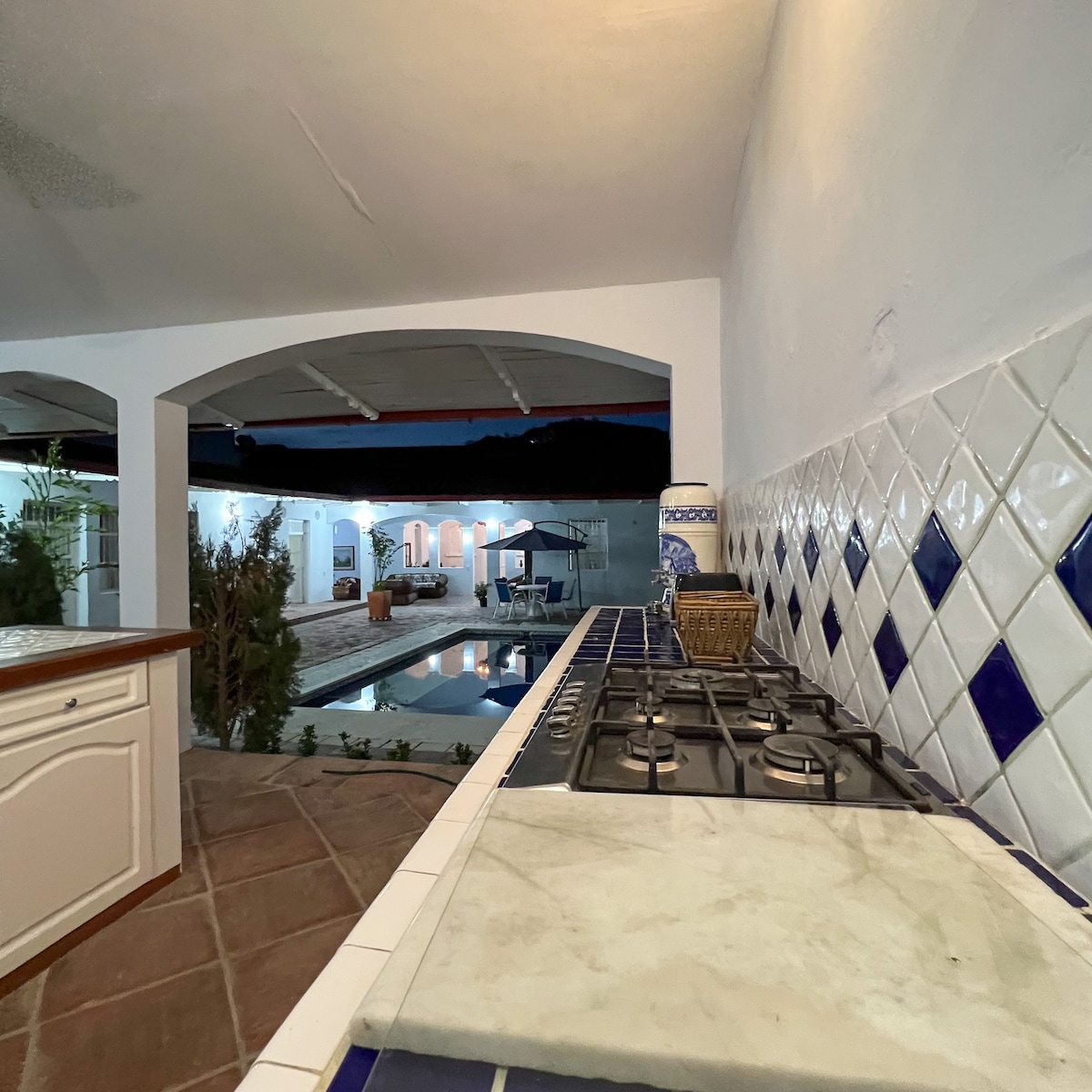 A kitchen area features a marble countertop with a gas stove, set against a backdrop of blue and white tiled walls. An open archway reveals a view of the pool area illuminated at night, with greenery visible around the outdoor space.