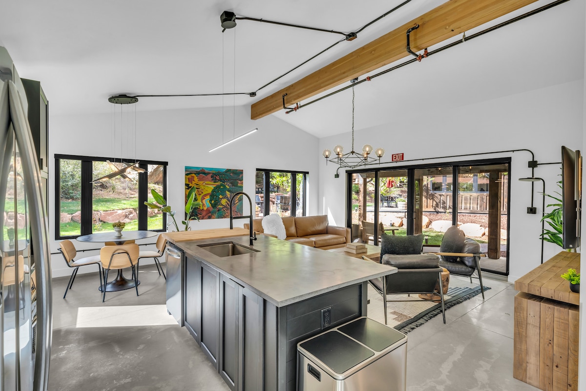 An open-concept living area features a modern kitchen island with a sleek design, surrounded by dining chairs. Large windows provide ample natural light and showcase views of the outdoor surroundings. A comfortable seating arrangement is situated near the wall-mounted television.