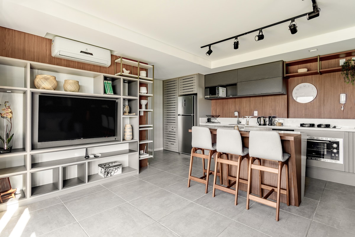 A modern kitchen and living area feature sleek cabinetry and a large wall-mounted television. Counter seating with three stools is positioned near the kitchen island, which includes an electric stove and built-in oven. Natural light fills the space through large windows, illuminating the neutral-toned decor.