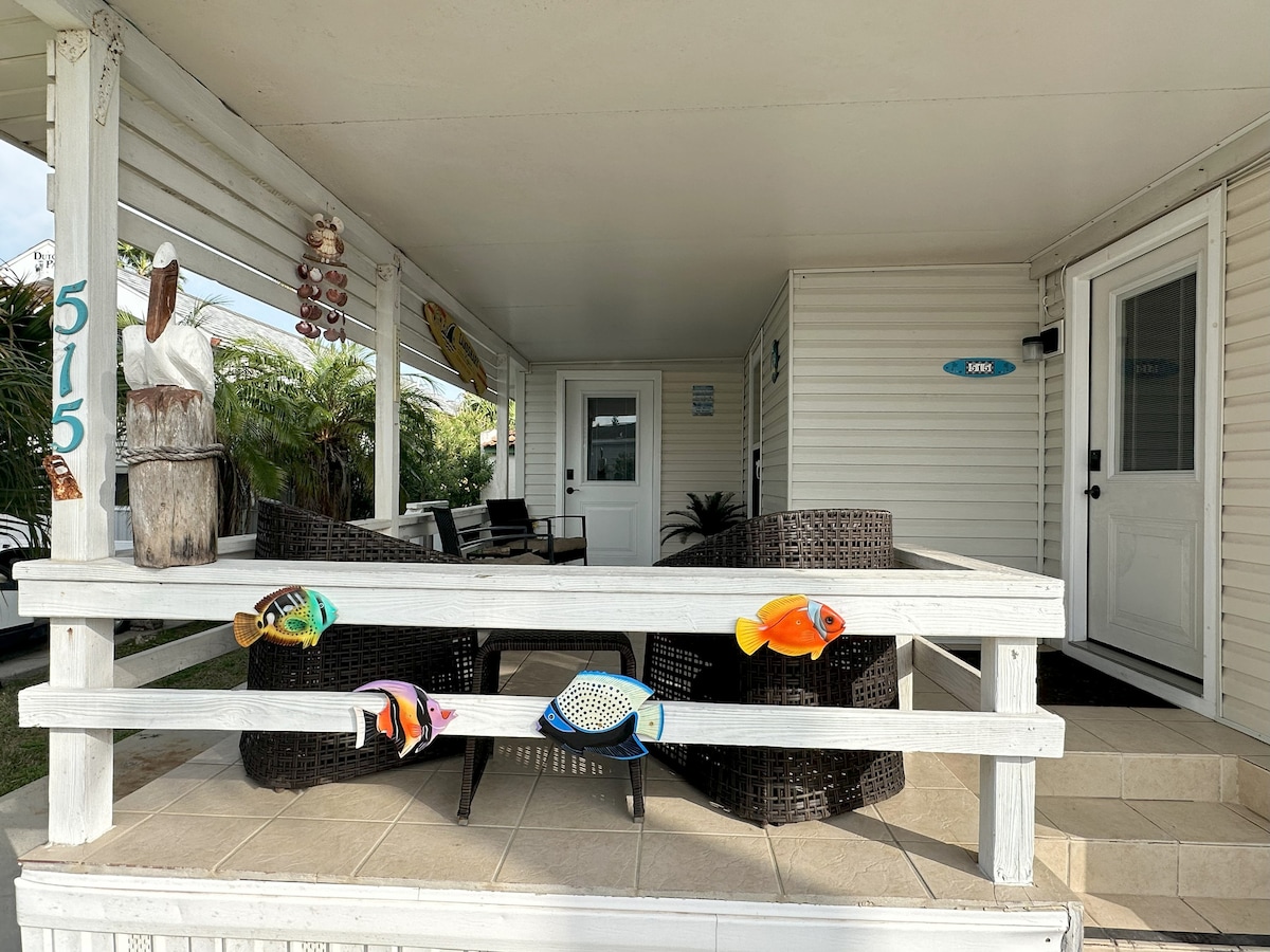 A covered patio features dark wicker lounge chairs arranged around a small table, inviting relaxation. Colorful fish decorations add a whimsical touch along the railing. The entrance door is visible on the right, with a welcoming atmosphere created by the surrounding greenery.