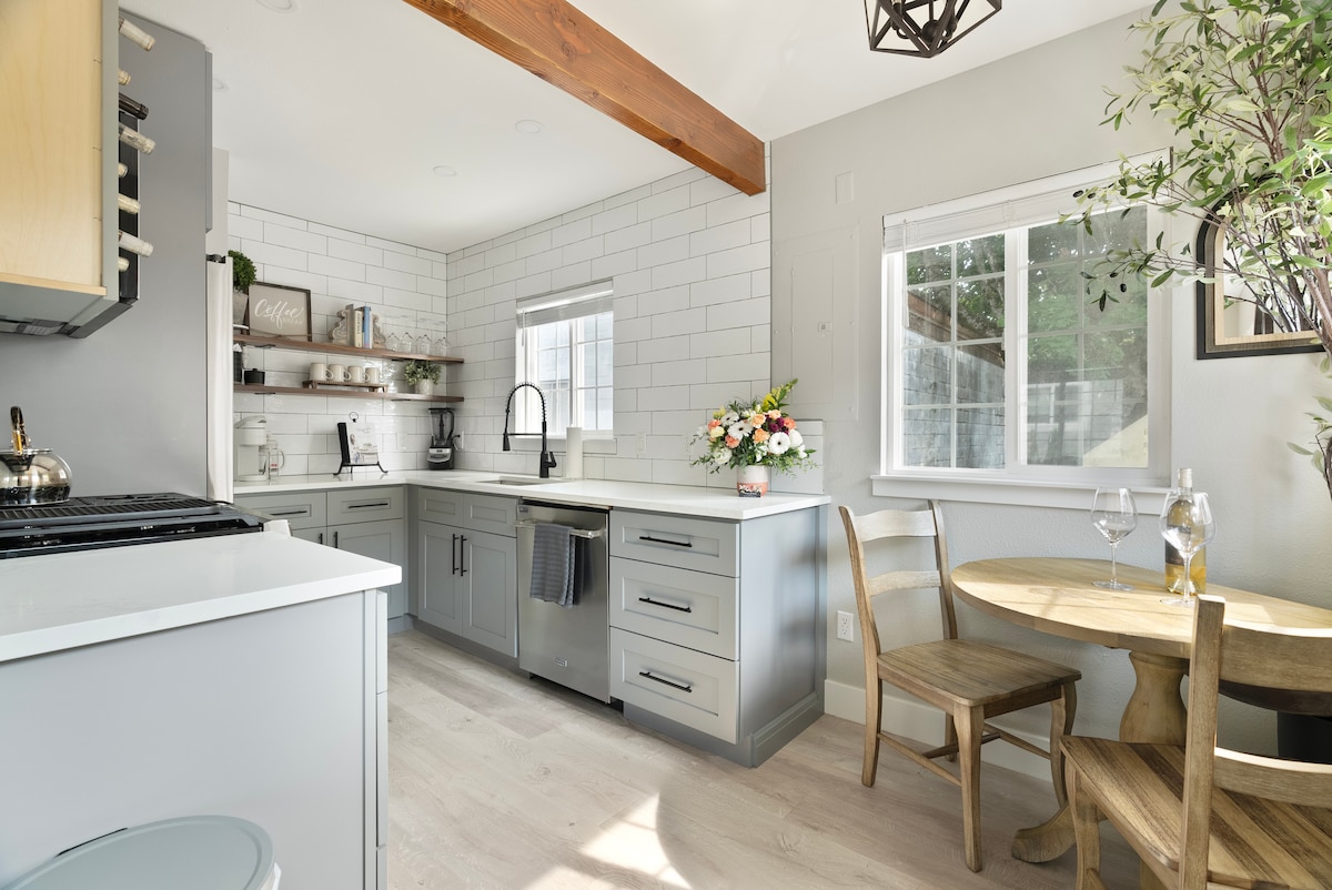 A modern kitchen is featured, equipped with stainless steel appliances and a spacious countertop. White tile backsplash complements the cabinetry, illuminated by natural light from the nearby window. A small dining nook with wooden chairs and a round table is positioned to the side, adorned with fresh flowers.