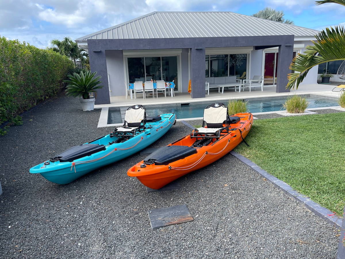 Two kayaks, one in bright blue and the other in vibrant orange, are positioned on a gravel surface near a modern, single-story house. The house features a covered terrace with a dining table and large windows, overlooking a pool surrounded by lush greenery.